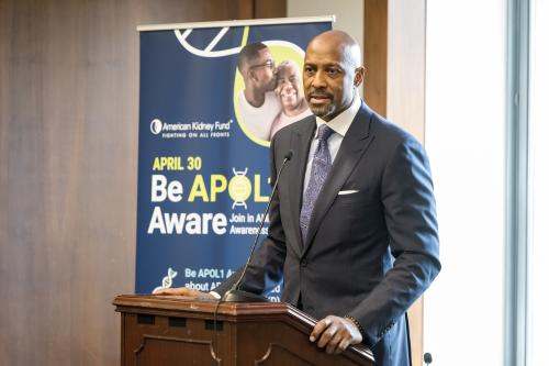 Alonzo Mourning speaking at a podium with a poster behind him that says, "Be APOL1 Aware"