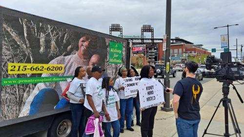 A group of women and children holding signs being interviewed by a camera crew in front of a billboard truck