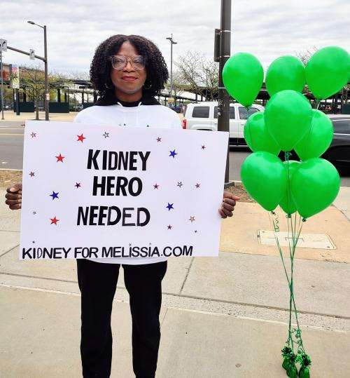 Melissia Baker standing next to green balloons holding a sign that reads "kidney hero needed kidneyformelissia.com"