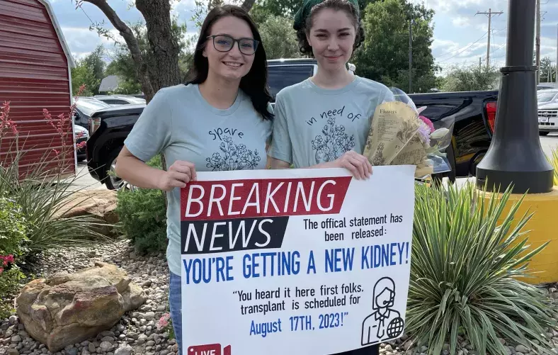 Savannah Stallbaumer and Katie Hallum holding a sign that reads, "Breaking News: you're getting a new kidney"