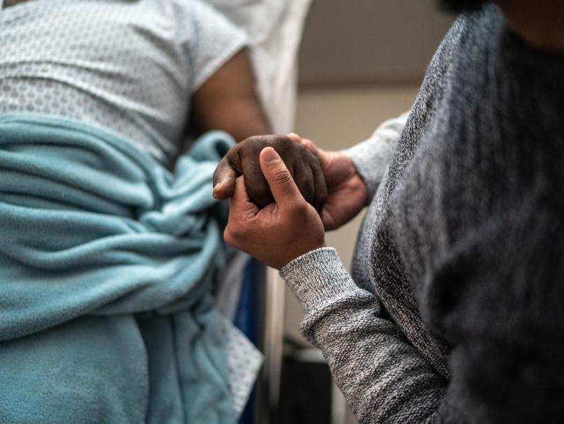 A person holding another person's hand while they lie in a hospital bed.